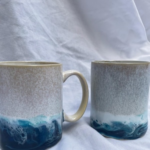 Puede incluir: Dos tazas de cerámica con una sección superior beige moteada que se transforma en una base vibrante de color azul océano y blanco. Las tazas tienen un acabado liso y brillante con un degradado de color, una taza incluye un asa curva.
