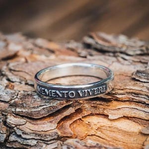 May include: A silver ring with the inscription "MEMENTO VIVERE" engraved around the band. The ring is set against a textured, brown wooden surface. The ring's design is simple and elegant.