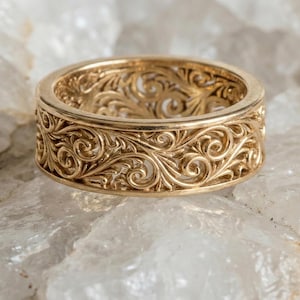 Puede incluir: Un anillo dorado con un diseño calado intrincado. El anillo presenta un patrón continuo de motivos florales y de volutas arremolinadas alrededor de la banda. El anillo está sobre un fondo claro y texturizado.