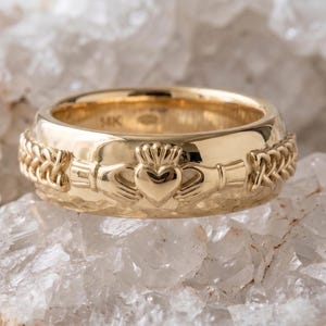 Puede incluir: Un anillo Claddagh dorado, con un diseño de corazón, corona y manos. El anillo está sobre un fondo texturizado blanco y gris. La banda del anillo tiene un diseño trenzado a cada lado del símbolo Claddagh.
