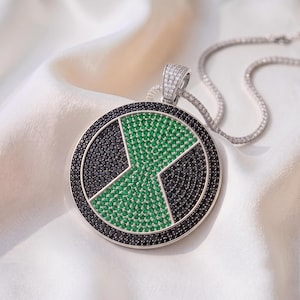 May include: A circular silver-toned pendant featuring a central green hourglass shape surrounded by black stones, all set within a round frame. The pendant is attached to a silver-toned chain, with both the bail and chain encrusted with small clear stones.