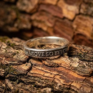 Może przedstawiać: Srebrny pierścień z wygrawerowanym napisem "AS ABOVE SO BELOW" na obwodzie. Pierścień umieszczony na tle brązowej, teksturowanej kory, podkreślający metaliczny połysk i szczegółowe grawerowanie.