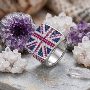 Puede incluir: Un anillo de plata con una cara cuadrada adornada con un diseño de la Union Jack. La bandera está creada con piedras preciosas azules, rojas y blancas. El anillo se presenta sobre un fondo de cristales y geodas de amatista.