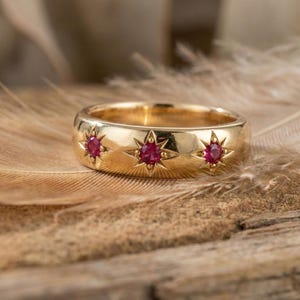 Peut inclure: Une bague en or ornée de trois motifs en forme d'éclat d'étoile, chacun orné d'une pierre précieuse rouge ronde. La bague est posée sur une plume et une surface en bois, créant une esthétique chaleureuse et naturelle.