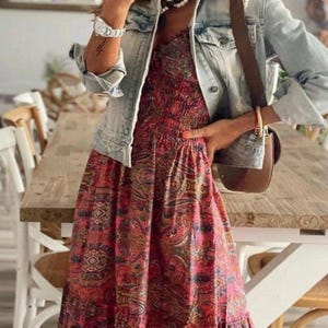 May include: A light blue denim jacket over a red and pink paisley print maxi dress. The dress has a fitted bodice and a flowing skirt. A brown leather shoulder bag is visible.