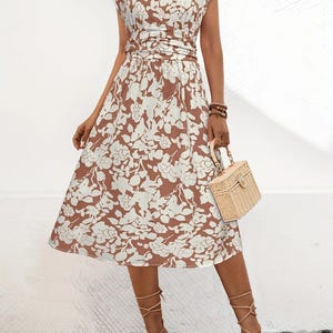 May include: A brown and white floral print dress with a V-neckline and cap sleeves. The dress has a cinched waist and a midi-length skirt. The model is wearing tan strappy heels and carrying a small woven handbag.