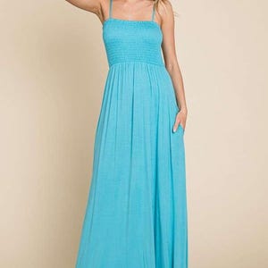 May include: A turquoise maxi dress with a smocked bodice and spaghetti straps. The dress features a flowing skirt and pockets. The model is wearing the dress and has her arm raised. The dress is made of a soft, lightweight fabric.