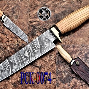 Könnte beinhalten: Ein Set aus drei Messern mit Holzgriffen und gemusterten Klingen. Das größte Messer hat eine Klinge von ca. 30 cm Länge. Das Set enthält eine Scheide für ein Messer. Der Text "PCK-0074" ist sichtbar.