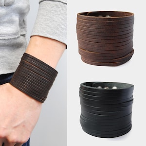 Op de afbeelding: Een donkerbruine leren manchetarmband wordt om een pols gedragen, met twee extra manchetten in de afbeelding. Eén is donkerbruin en de andere is zwart. De armbanden zijn gemaakt van meerdere horizontale leren stroken.