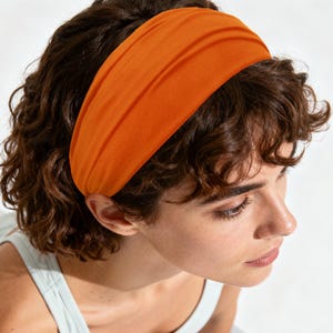 Può includere: Una fascia larga in tessuto arancione tinta unita indossata sulla fronte. La fascia presenta una consistenza liscia e opaca e un design semplice e privo di decorazioni che aderisce saldamente ai capelli, aggiungendo un tocco di colore.