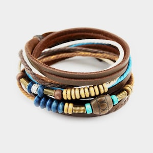 Könnte beinhalten: Ein mehrsträngiges Armband mit braunem Leder, weißen und türkisfarbenen Kordeln, Holzperlen, blauen Perlen und einem dekorativen Schildkrötenanhänger. Das Armband ist um sich selbst gewickelt und erzeugt einen geschichteten Look.