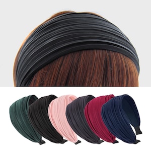 Könnte beinhalten: Ein schwarzes, plissiertes Stirnband auf braunem Haar. Darunter eine Auswahl an Stirnbändern in verschiedenen Farben: Grün, Schwarz, Rosa, Grau, Burgunderrot und Marineblau. Jedes Stirnband hat ein breites, plissiertes Design und ein schwarzes Gummiband.