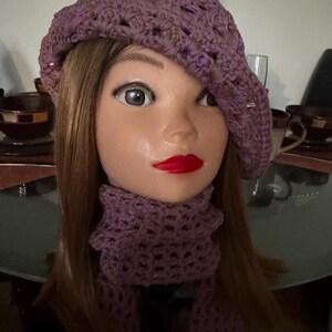May include: A lavender crochet beret and matching scarf set. The beret is worn on a mannequin head with brown hair and red lipstick. The scarf is a cowl style with a black bow. The set is displayed on a glass table.