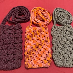 May include: Three handmade crochet phone bags in shades of purple, orange, and gray. Each bag has a long strap for wearing across the body. The bags are made with a textured stitch pattern, perfect for carrying a phone or small items.