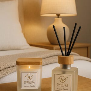 Op de afbeelding: Een brandende aromakaars en een rietverspreider op een houten oppervlak. De kaars is vierkant met een houten deksel en de diffuser heeft zwarte rietjes. Beide hebben het label "MONICA VALADARES Aroma Candle."