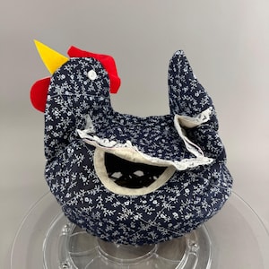May include: A quilted fabric chicken-shaped tea cozy featuring a dark navy blue background with a white floral pattern. The design includes a bright yellow beak, a red comb, a white button eye, and a decorative white lace trim around the opening.