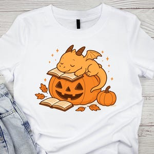 May include: White t-shirt with a cartoon dragon sleeping on a carved pumpkin. The dragon is orange with brown horns and wings. The pumpkin has a carved face and is surrounded by books and autumn leaves. A small pumpkin is to the side.