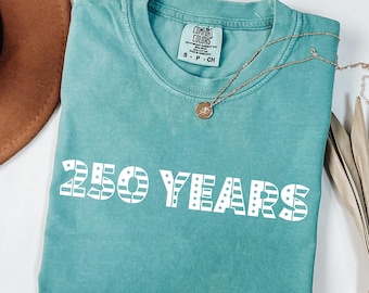 May include: A teal Comfort Colors t-shirt with the text "250 YEARS" in white block letters. A gold necklace with a pendant is draped across the shirt. The shirt is folded, and a brown hat is visible.