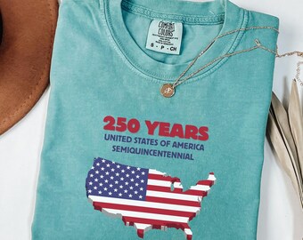 May include: A turquoise Comfort Colors t-shirt with the text "250 YEARS UNITED STATES OF AMERICA SEMIQUICENTENNIAL" in red and white. The shirt features a 3D map of the United States with the American flag design. A gold necklace is draped across the shirt.