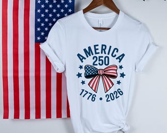 May include: White t-shirt with a patriotic design. The shirt features the text "AMERICA 250" in blue, along with a bow in red, white, and blue. The shirt also includes the years "1776" and "2026" and is hanging in front of an American flag.