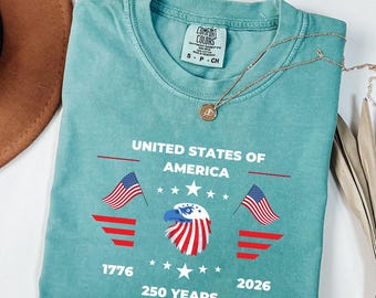 May include: A teal Comfort Colors t-shirt with the text "UNITED STATES OF AMERICA" and the years "1776" and "2026". The design includes American flags, stars, and an eagle graphic. A gold necklace is draped across the shirt.