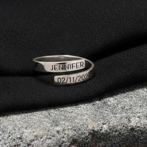 May include: A silver wrap ring engraved with the name "JENNIFER" and the date "02/11/2023". The ring is shown on a textured grey stone, set against a black fabric backdrop.