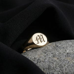 May include: A gold signet ring with an oval face, engraved with the letter "M" in an elegant, stylized font. The ring is set on a grey stone, with a black fabric in the background.