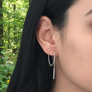 May include: A close-up of a person's ear adorned with two silver earrings. One is a small stud, and the other is a chain earring with a dangling bar. The earrings are set against a backdrop of green foliage.
