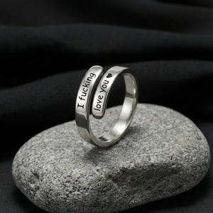 May include: A silver-colored ring with the words "I fucking love you" engraved on it. The ring has a wrap-around design, with the text split across the two bands. A small heart symbol follows the words "love you".
