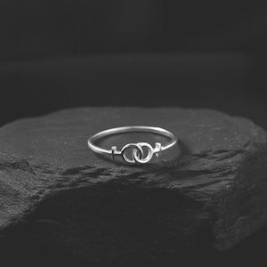 May include: A silver ring featuring a design of two interlocking circles, connected by a thin band. The ring is set against a dark, textured background, highlighting its delicate details and metallic sheen.