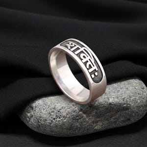 May include: A silver ring with engraved text in a stylized font. The ring is displayed on a grey stone against a black fabric backdrop. The ring's design features a band with the engraved text.