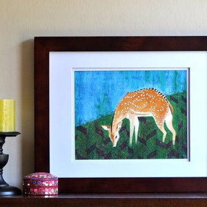 May include: A framed print of a watercolor painting depicting a spotted deer grazing in a green field with a blue sky in the background.