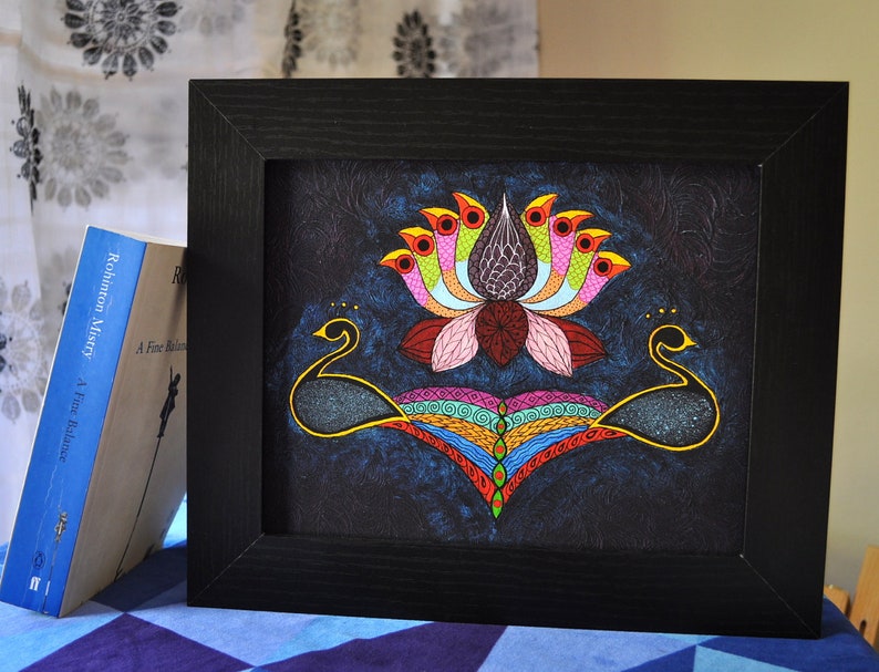 May include: A framed painting of a colorful lotus flower with peacocks on either side. The lotus has pink petals and a dark purple center. The peacocks are black with gold accents. The background is a dark blue.