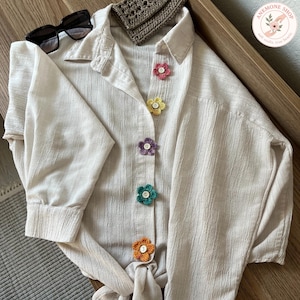 Può includere: Una camicia color crema con bottoni a forma di fiori all'uncinetto colorati. La camicia è abbinata a occhiali da sole e a un piccolo portafoglio all'uncinetto marrone. La camicia è annodata in vita.