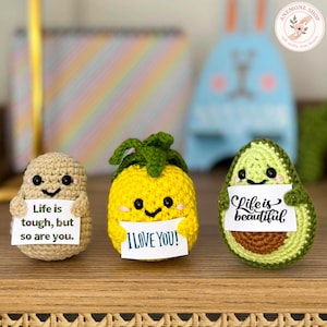 Puede incluir: Tres figuras de comida de ganchillo: una patata, una piña y un aguacate. La patata sostiene un cartel que dice "Life is tough, but so are you." La piña dice "I LOVE YOU!" y el aguacate dice "Life is beautiful."