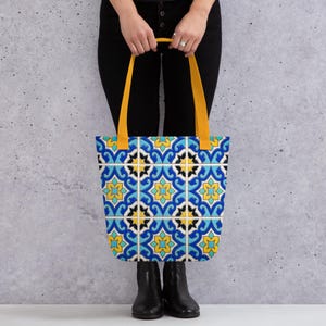 May include: A tote bag featuring a repeating pattern of blue, yellow, and white geometric tile designs. The bag has two solid mustard yellow shoulder straps and a rectangular body with a vibrant, symmetrical floral-inspired motif throughout.
