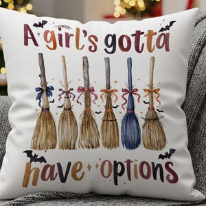 May include: White decorative pillow with the text "A girl's gotta have options" and illustrations of six brooms with colorful bows. The brooms are brown, with varying colors of blue, pink, and orange bows. Black bats are scattered around the text.