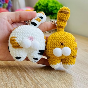 以下が含まれることがあります： 2つの手編みの猫のお尻のキーホルダー。1つは白地に黒と茶色の縞模様、もう1つは黄色の地に茶色の縞模様です。どちらも白いふわふわのお尻で、シルバーのキーリングが付いています。キーホルダーは手作りです。