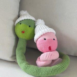 Può includere: Due giocattoli a forma di serpente all'uncinetto, uno verde e uno rosa. Entrambi hanno cappelli bianchi con pompon, occhi neri e bocca rossa. Il corpo del serpente verde forma un anello attorno al serpente rosa.