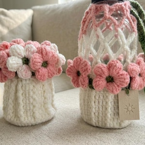May include: Two handmade crochet items: a small vase and a water bottle holder. Both are white with pink and white flower accents. The bottle holder has a green strap. The vase is filled with pink and white flowers.
