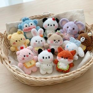 Può includere: Una collezione di portachiavi a forma di animale all'uncinetto fatti a mano in un cestino di vimini. I portachiavi presentano vari animali, tra cui un panda, un orso, una volpe e un elefante, in colori pastello. Ogni portachiavi ha un anello di metallo.