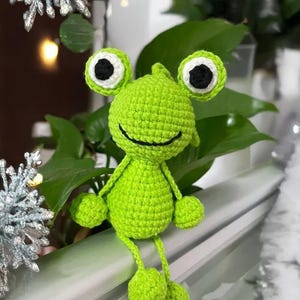 May include: A handmade, bright green crochet frog doll with large, round eyes and a smiling mouth. The frog has small arms and legs with round feet. The frog is sitting in front of green leaves.