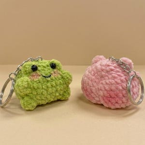 May include: Two crochet keychains: a green frog with black eyes and pink cheeks, and a pink, round creature. Both have silver key rings attached. The frog keychain is approximately 2.5 inches wide.