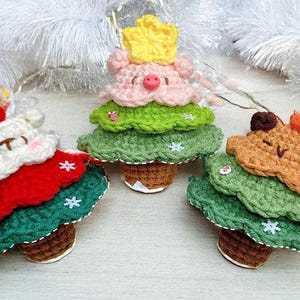 May include: Three handmade crochet Christmas tree ornaments. One is white and red, one is pink with a yellow crown, and the other is brown. Each has a brown base and green layers, with white snowflake accents.