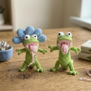 May include: Two handmade crochet lizards in light green yarn with pink tongues and wide eyes. One lizard wears a large, light blue crocheted flower on its head. Both figures have open mouths and are standing in an upright, arms-outstretched pose.