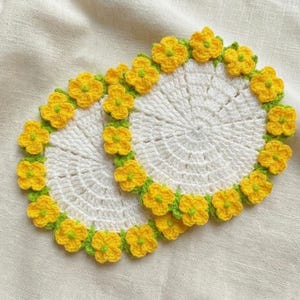 May include: Two handmade crochet coasters. Each coaster is white with a yellow flower border and green leaves. The coasters are circular and sit on a cream-colored fabric.