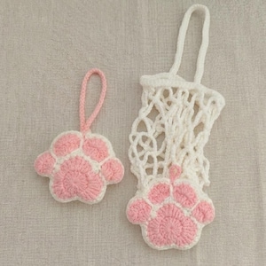 May include: Two handmade crochet items: a pink and white paw print ornament with a loop and a white net bag with a pink paw print base. The items are made of yarn and are on a neutral background.