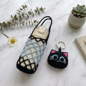 May include: A beige water bottle in a black and light blue crochet carrier, alongside a black cat-shaped keyring with pink details. A small succulent in a white pot and a notebook with a pencil are also present.