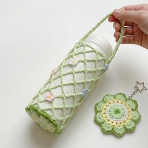 May include: A light green crocheted water bottle holder with a floral pattern, holding a white bottle. The holder has a handle and is decorated with small pink and blue flowers. A matching coaster and keychain are also present.