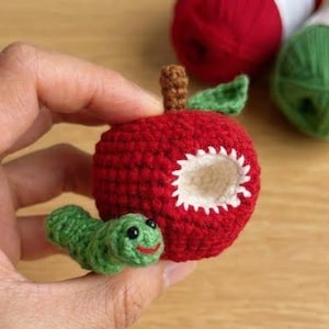 May include: A small red crocheted apple with a brown stem and a green leaf, featuring a white bite mark on the side. A small green crocheted caterpillar with black eyes and a red stitched mouth is attached to the base of the red apple.